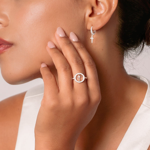 Close-up of a christian woman wearing a sterling silver compass ring and sparkly cross hoop earring on a neutral background from Rizen Jewelry's Holiday Collection.