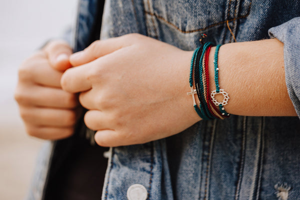 Rizen Jewelry Made 4 Ministries bracelet stack on model wearing jean jacket. Cross, crown of thorns bracelets in azure blue with multi cord colorful christian friendship bracelet. 