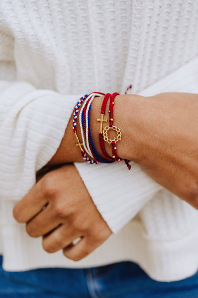 Christian model wearing gold cross bracelet engraved "STAY SALTY" hand braided garnet red cotton cords by Rizen Jewelry Made 4 Ministries collection. Layered with beaded crown of thorns, and red, white, blue, we trust in god, and multi cord bracelets.  