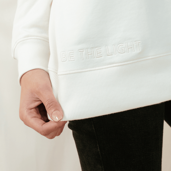 Close up "BE THE LIGHT" embroidery of the sand off white "Chosen. Ephesians 1:4" Peruvian cotton crew sweatshirt from the Be The Light Collection by Rizen Jewelry.